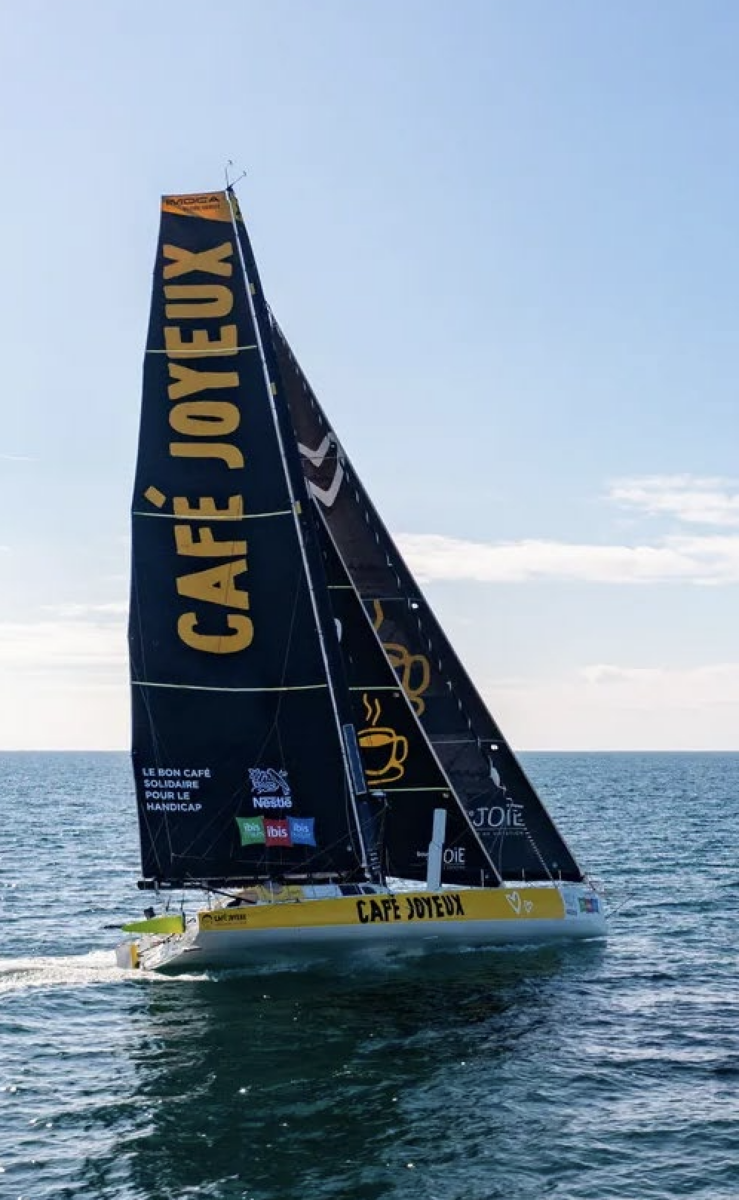 Un voilier jaune et noir arborant Café Joyeux et 2 Mains 4 Cornes sur ses voiles glisse en haute mer sous un ciel dégagé. Le bateau porte les logos des sponsors et des tasses de café stylisées. - Édition Offset 5 (Partenaire de votre communication locale en Vendée)
