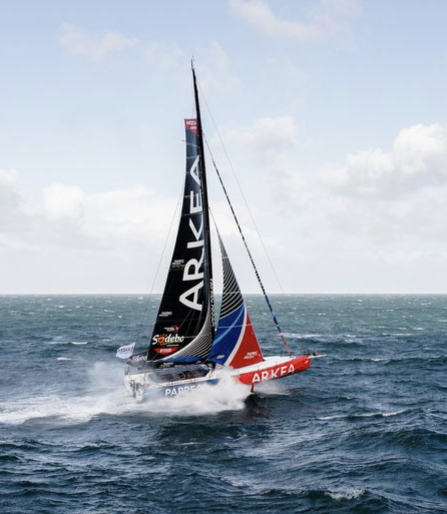 Un voilier de course arborant la marque ARKEA sur sa voile et l'emblème 2 Mains 4 Cornes fend les vagues agitées de l'océan, créant des embruns sous un ciel partiellement nuageux. Le bateau arbore des couleurs vives : rouge, noir et bleu. - Édition Offset 5 (Partenaire de votre communication locale en Vendée)