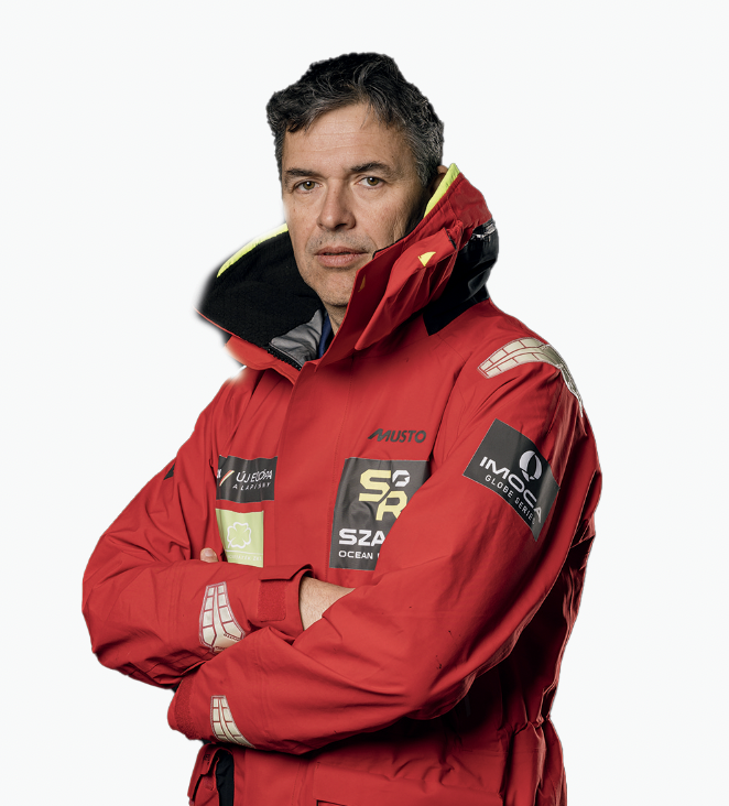 Un homme aux cheveux bruns courts portant une veste de voile rouge avec plusieurs logos de sponsors, dont 2 Mains 4 Cornes, se tient debout, les bras croisés, regardant la caméra avec assurance sur un fond blanc. - Édition Offset 5 (Partenaire de votre communication locale en Vendée)