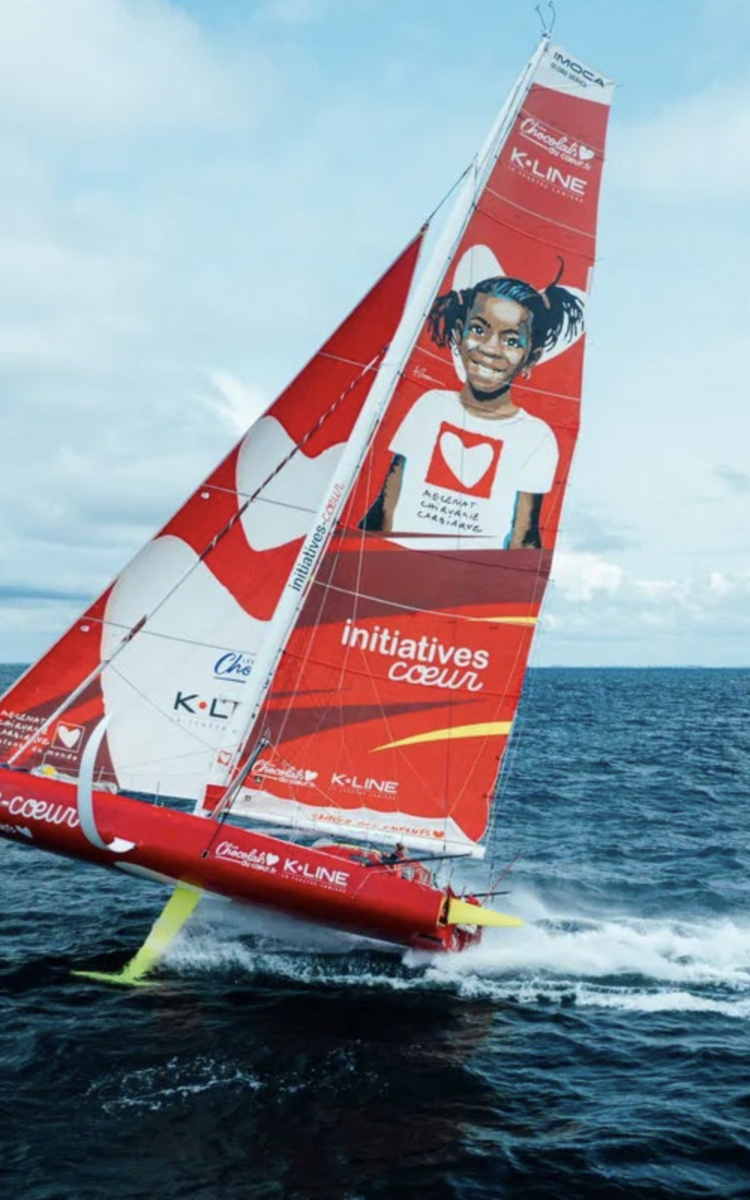 Un voilier rouge et blanc portant l'inscription 2 Mains 4 Cornes, avec une grande image d'un enfant souriant et un logo en forme de cœur sur la voile, navigue à vive allure sur l'océan bleu sous un ciel nuageux. - Édition Offset 5 (Partenaire de votre communication locale en Vendée)