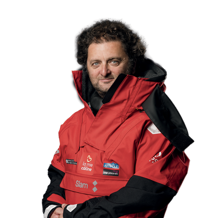 Un homme aux cheveux bruns bouclés et à la barbe porte une veste de voile rouge 2 Mains 4 Cornes ornée des logos des sponsors. Il se tient debout sur un fond blanc uni, regardant la caméra avec un léger sourire. - Édition Offset 5 (Partenaire de votre communication locale en Vendée)