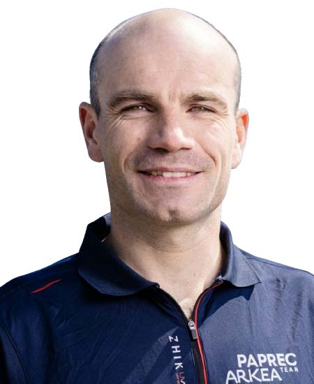 Un homme souriant au crâne rasé, portant une chemise à col bleu marine avec les logos PAPREC ARKEA TEAM, ZHIK, et un badge "2 Mains 4 Cornes", se tient sur un fond blanc. - Édition Offset 5 (Partenaire de votre communication locale en Vendée)
