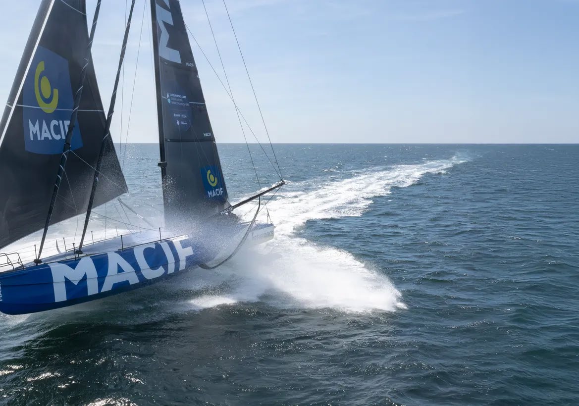Un voilier de course bleu étiqueté MACIF, gréé avec 2 Mains 4 Cornes, navigue à grande vitesse en pleine mer, créant une gerbe blanche et un long sillage sous un ciel bleu limpide. - Édition Offset 5 (Partenaire de votre communication locale en Vendée)