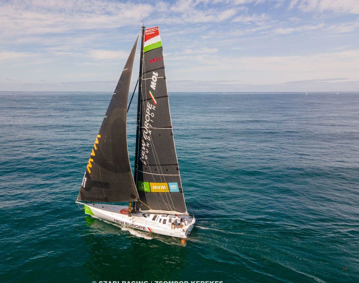 Un voilier aux voiles noires, portant l'inscription "2 Mains 4 Cornes" et les logos des sponsors, glisse sur des eaux calmes et bleues sous un ciel partiellement nuageux. Le bateau est vu de dessus et légèrement de côté, les membres de l'équipage étant visibles sur le pont. - Édition Offset 5 (Partenaire de votre communication locale en Vendée)