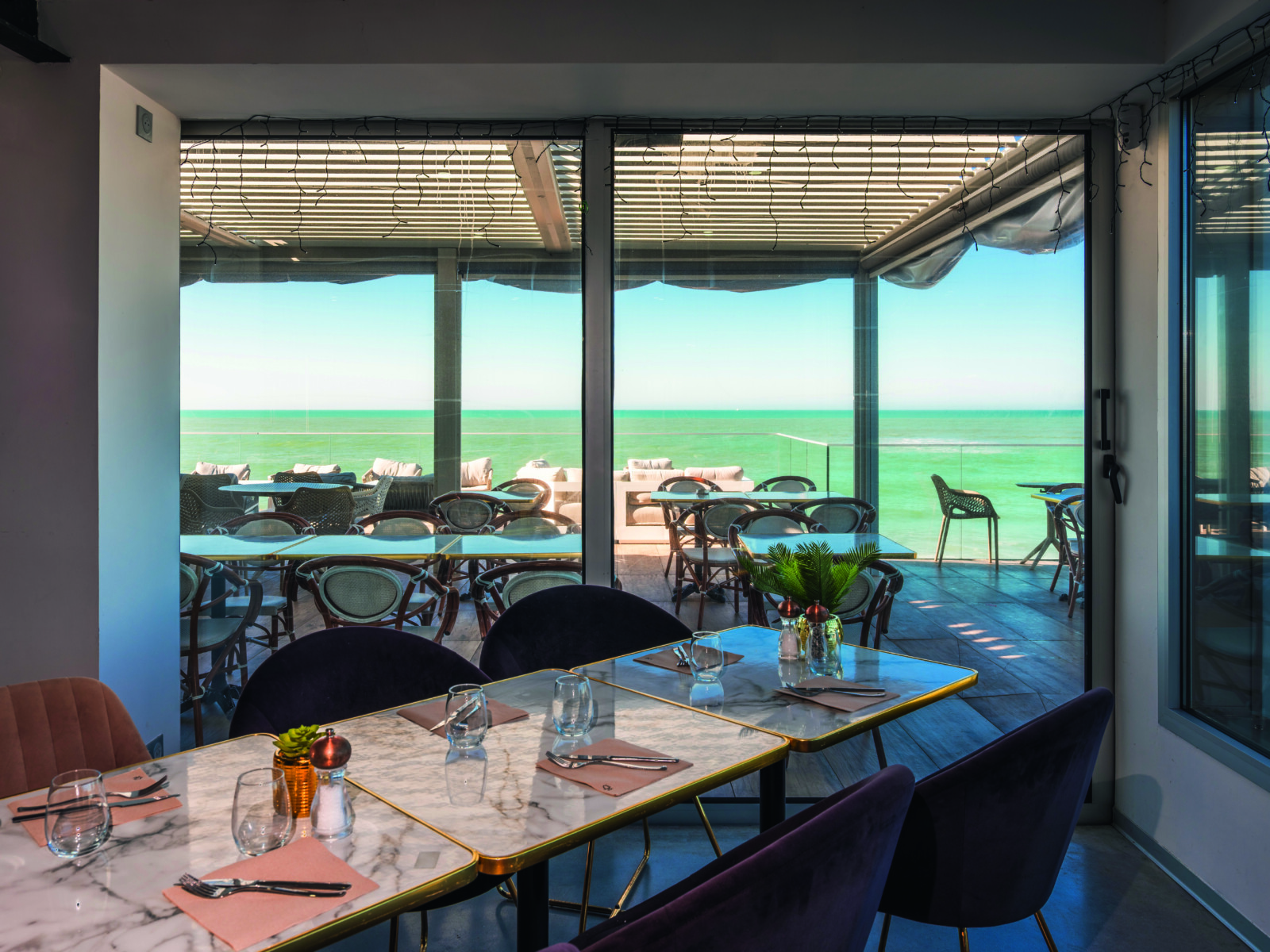 Le Bouchon d'Or, un restaurant moderne, dispose de tables en marbre, donnant sur un patio extérieur ensoleillé avec des chaises et des tables, et offre une vue dégagée sur la mer turquoise à travers de grandes baies vitrées. - Édition Offset 5 (Partenaire de votre communication locale en Vendée)
