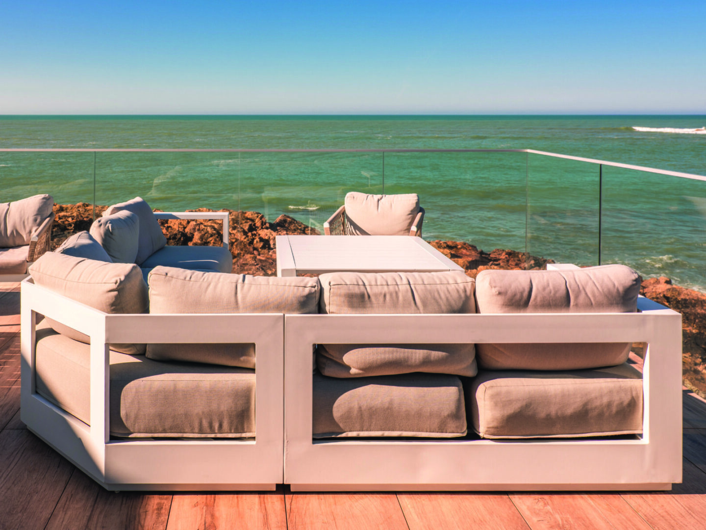Terrasse moderne du Bouchon d'Or avec des canapés et une table gris clair, donnant sur un rivage rocheux et un océan turquoise sous un ciel bleu clair, entourée d'une balustrade en verre. - Édition Offset 5 (Partenaire de votre communication locale en Vendée)
