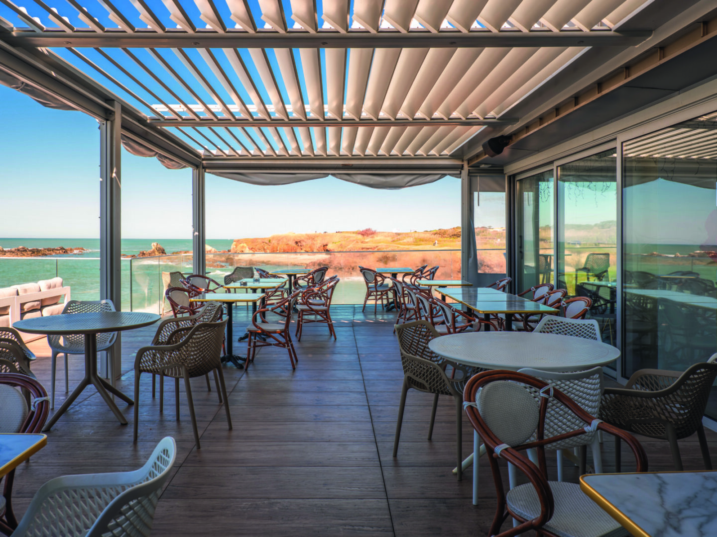 La terrasse du Bouchon d'Or se compose de tables et de chaises vides sous un toit en lattes, avec vue sur une mer calme et un rivage rocheux lointain par une journée claire et ensoleillée. - Édition Offset 5 (Partenaire de votre communication locale en Vendée)