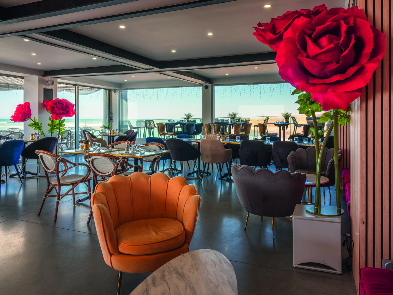 Le Bouchon d'Or est un restaurant moderne et lumineux, avec des chaises en velours coloré, des tables en verre et de grandes roses rouges décoratives. Des fenêtres allant du sol au plafond offrent une vue sur la terrasse extérieure et la mer. - Édition Offset 5 (Partenaire de votre communication locale en Vendée)