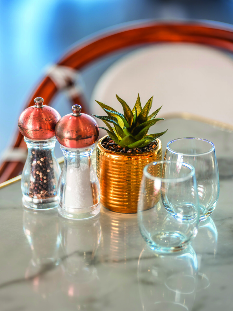 Une petite plante succulente en pot, des salières et poivrières à couvercle en cuivre et deux tasses en verre vides sont disposées sur une table en marbre au Bouchon d'Or, avec une chaise floue à l'arrière-plan. - Édition Offset 5 (Partenaire de votre communication locale en Vendée)