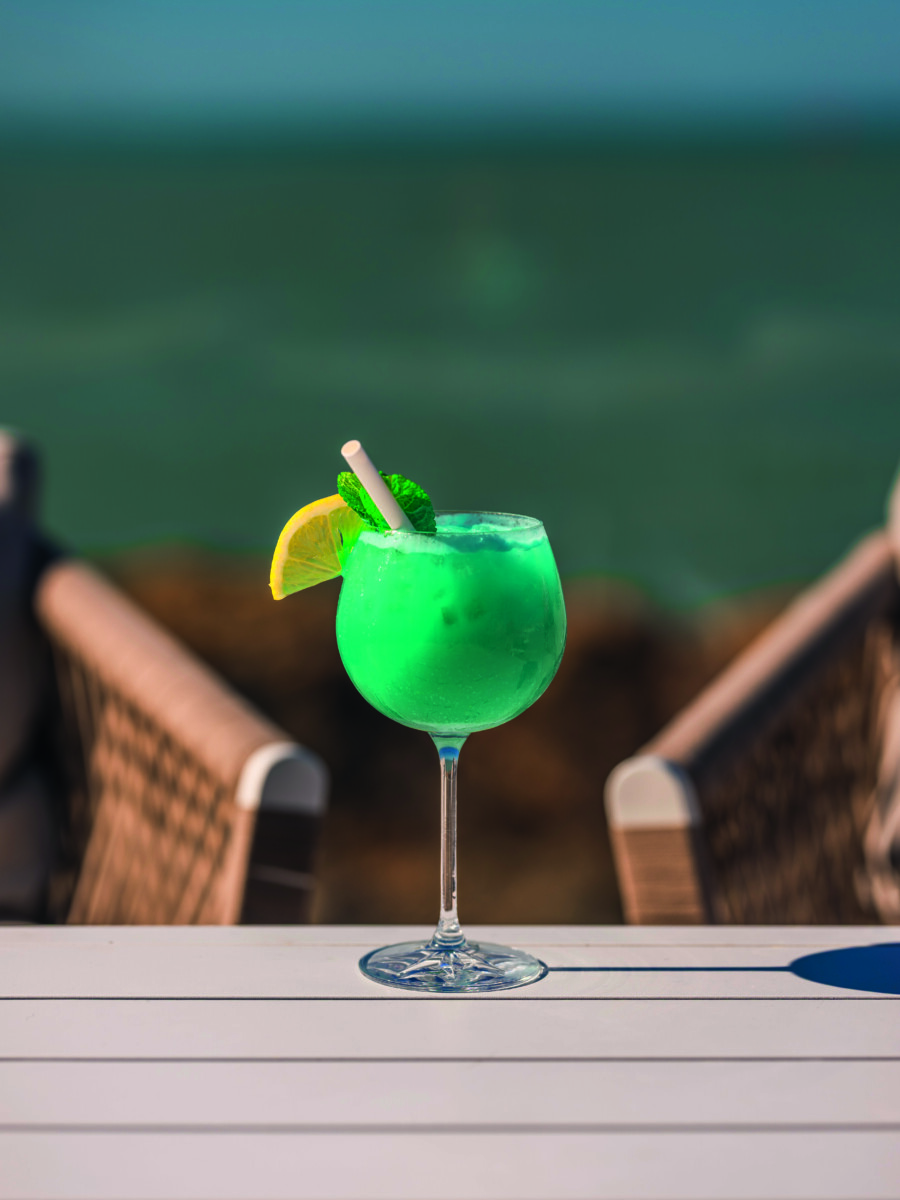 Un cocktail vert vif du Bouchon d'Or, garni d'une rondelle de citron et de menthe, est posé sur une table blanche à l'extérieur, avec deux chaises en osier et de l'eau floue en arrière-plan. - Édition Offset 5 (Partenaire de votre communication locale en Vendée)