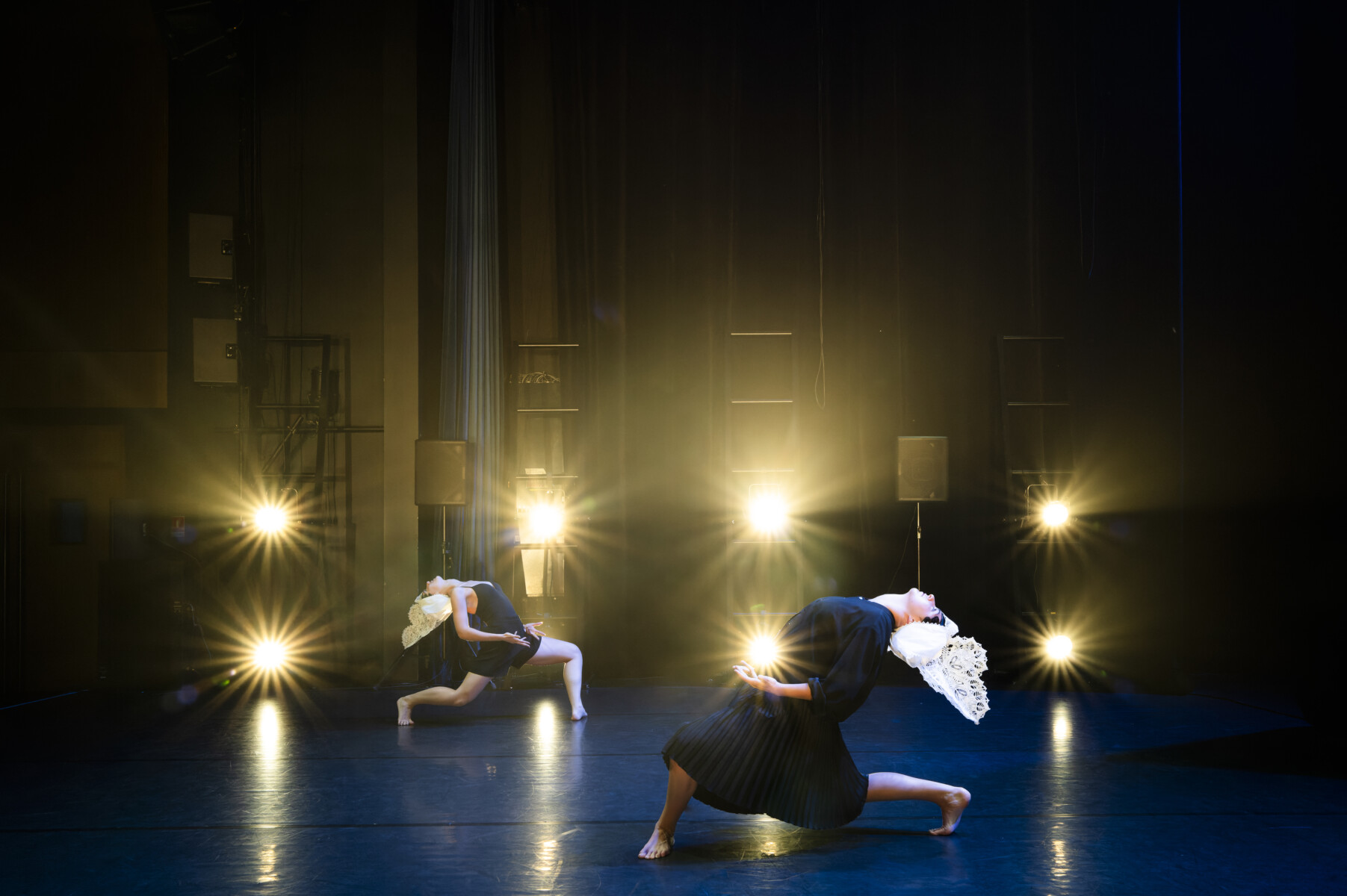 Sur une scène faiblement éclairée, deux danseurs prennent une pose dramatique, tous deux vêtus de costumes noirs et de coiffes blanches. Les lumières de la scène brillent derrière eux, projetant de longues ombres qui rappellent l'énergie frappante que l'on trouve au Bike Park montaigu-Vendée. - Édition Offset 5 (Partenaire de votre communication locale en Vendée)