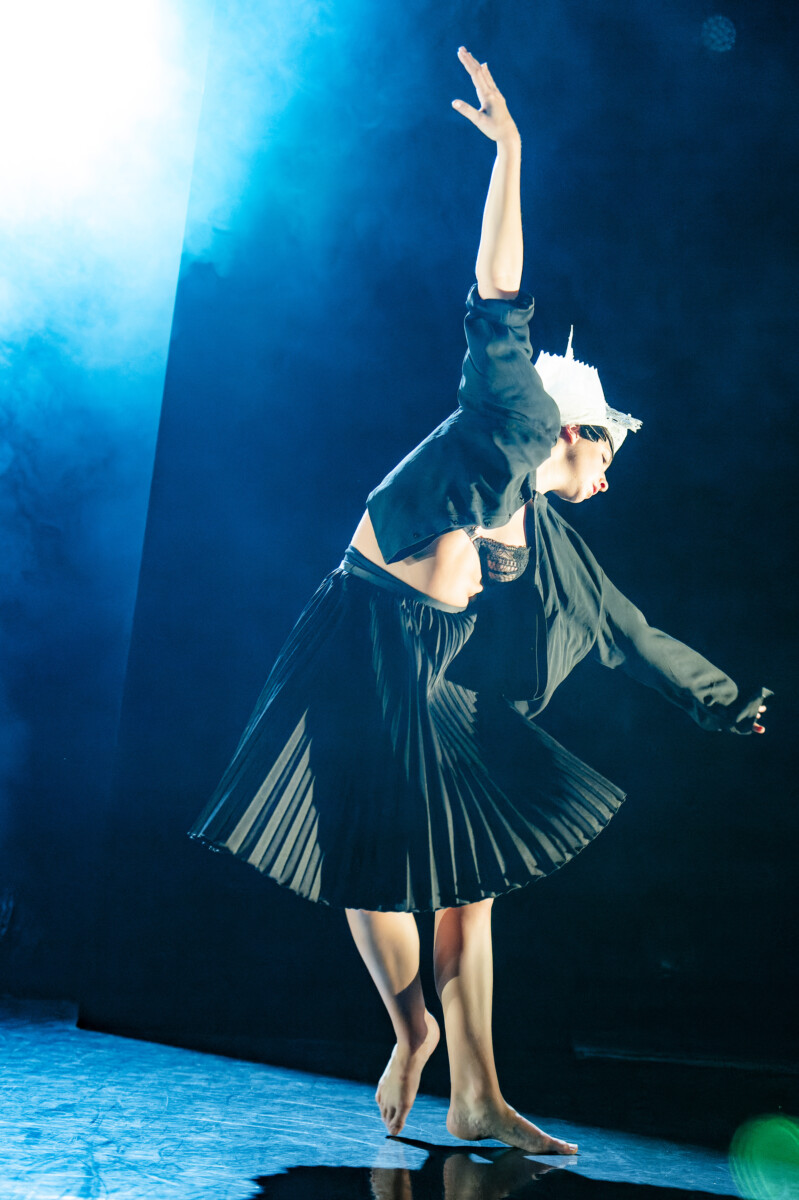 Une danseuse aux pieds nus, vêtue d'une jupe plissée noire, d'une veste courte et d'une couronne blanche, pose gracieusement sur scène sous un éclairage bleu. Son élégance rappelle celle d'un spectacle au Bike Park Montaigu-Vendée, un bras levé et l'autre tendu vers le bas. - Édition Offset 5 (Partenaire de votre communication locale en Vendée)