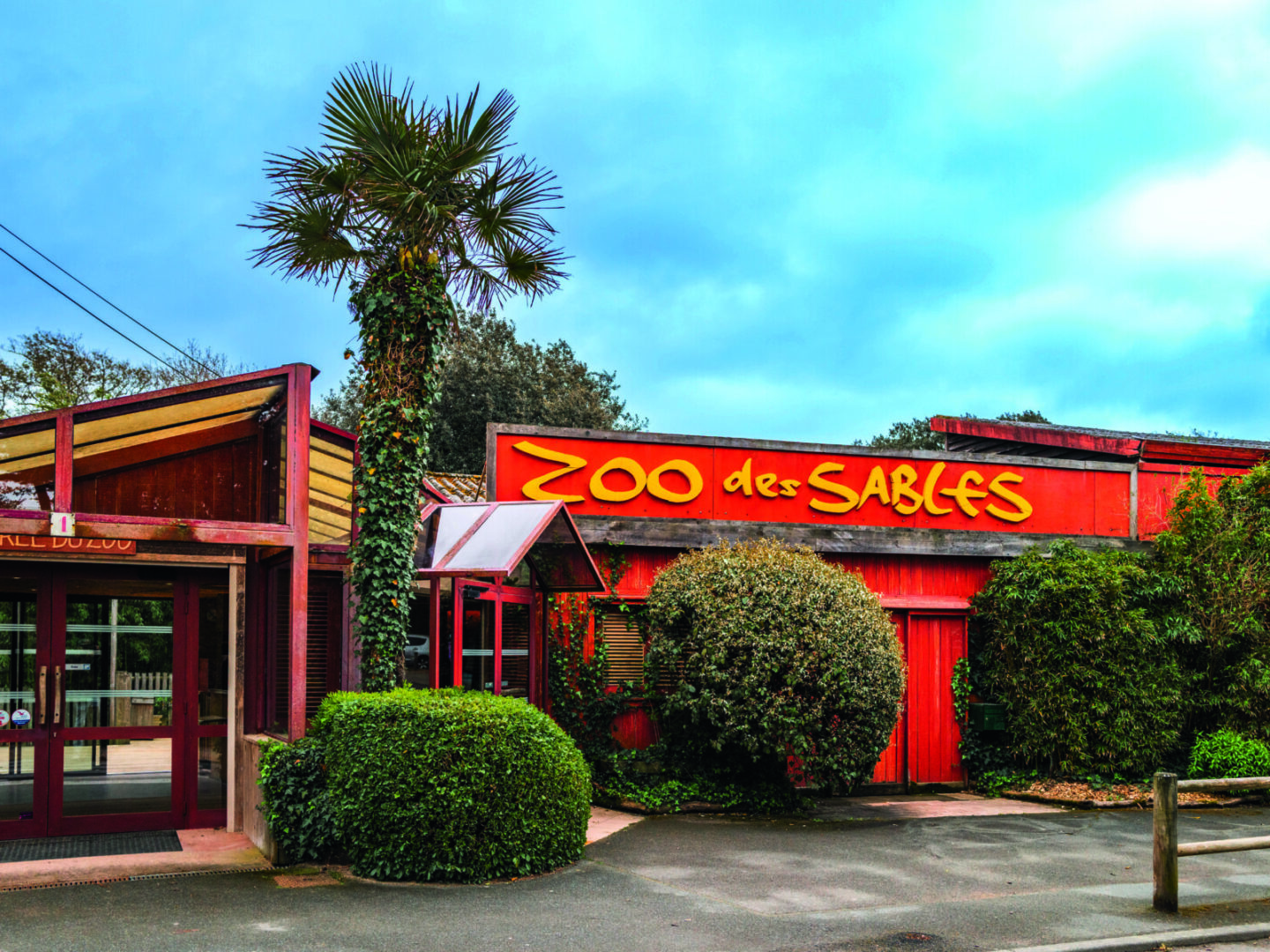L'image montre l'entrée du Zoo des Sables, avec un bâtiment rouge, un panneau jaune, des buissons verts et un grand palmier sous un ciel bleu partiellement nuageux. Tout près se trouve l'aire d'activité 2 Mains 4 Cornes pour les enfants. - Édition Offset 5 (Partenaire de votre communication locale en Vendée)