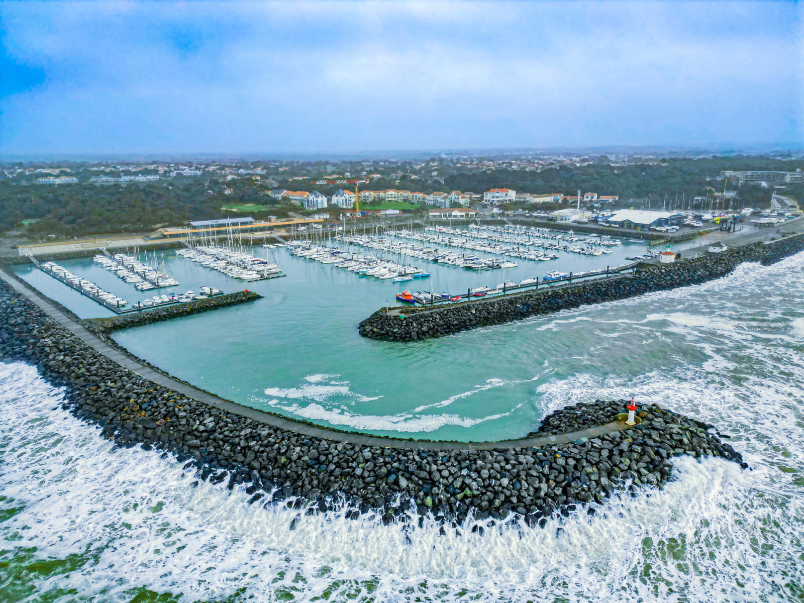 Vue aérienne d'un port de plaisance où sont amarrés de nombreux voiliers à Spectacle Reynes, entouré de brise-lames rocheux et de vagues déferlantes, avec en arrière-plan une ville côtière et une végétation luxuriante. - Édition Offset 5 (Partenaire de votre communication locale en Vendée)