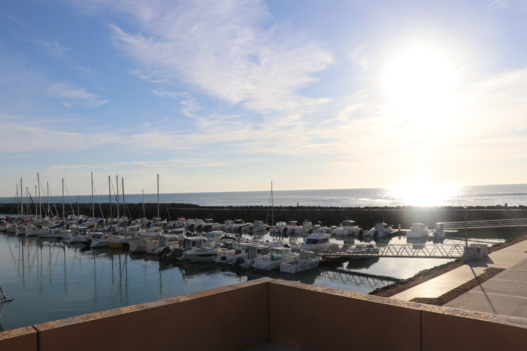 Une marina à Spectacle Reynes, avec de nombreux bateaux amarrés, des eaux calmes et un brise-lames en pierre, est photographiée au coucher du soleil, alors que le soleil est bas dans le ciel et que de légers nuages parsèment l'horizon au-dessus de l'océan. - Édition Offset 5 (Partenaire de votre communication locale en Vendée)