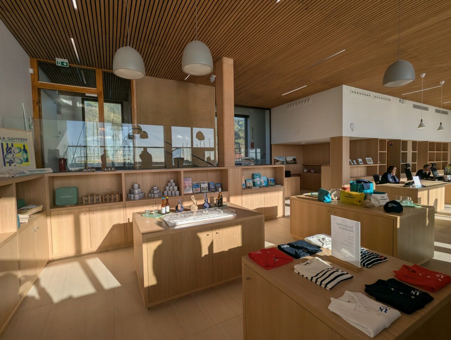 Une boutique de cadeaux moderne, dotée d'étagères en bois et de tables d'exposition sur lesquelles sont présentés des livres, des vêtements et des souvenirs de Spectacle Reynes. De grandes baies vitrées laissent entrer la lumière du soleil, et deux personnes travaillent derrière le comptoir. L'endroit dégage une atmosphère chaleureuse et accueillante. - Édition Offset 5 (Partenaire de votre communication locale en Vendée)